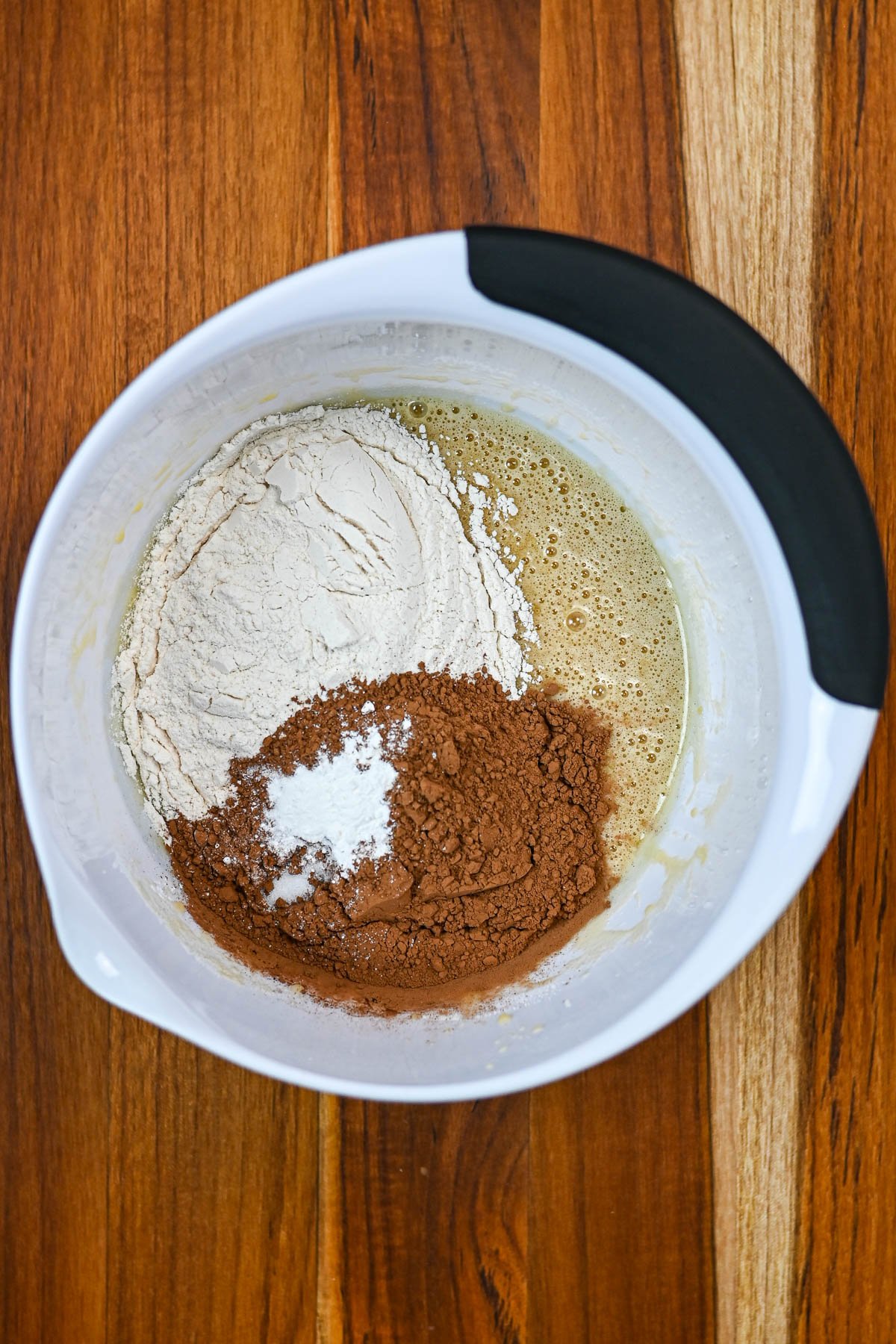 White mixing bowl with flour, cocoa powder, and baking soda on a wooden surface, ready to be mixed for delicious Kiss Crinkle Cookies or classic chocolate crinkles.