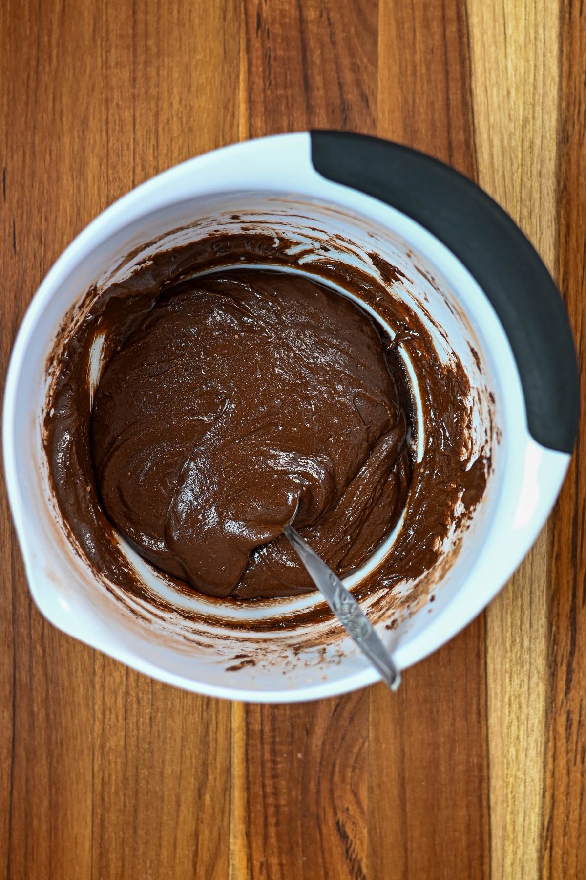A white bowl with chocolate batter for Kiss Crinkle Cookies and a spoon, placed on a wooden surface.