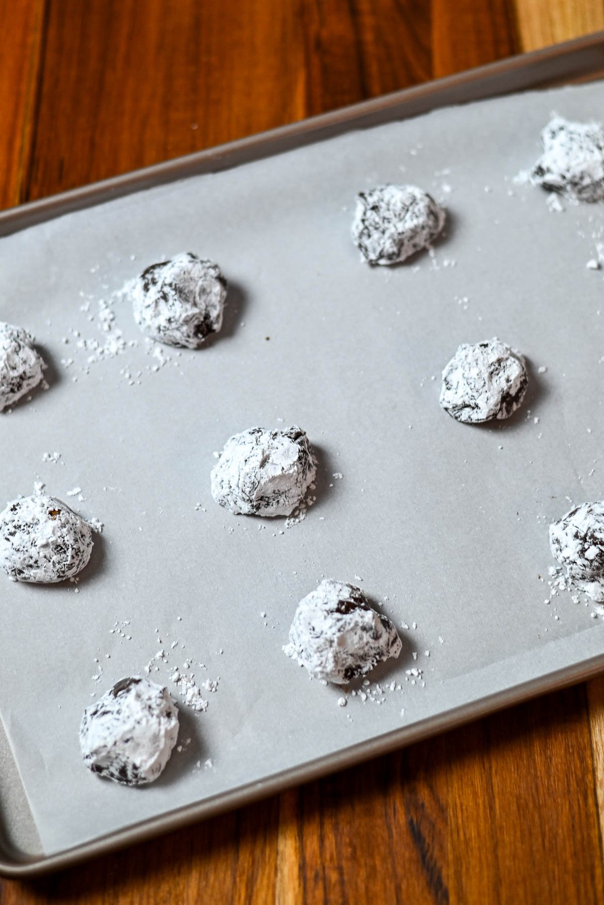 Chocolate crinkle cookie dough balls coated in powdered sugar on a baking sheet lined with parchment paper-these chocolate crinkles promise fudgy centers and classic crackled tops.