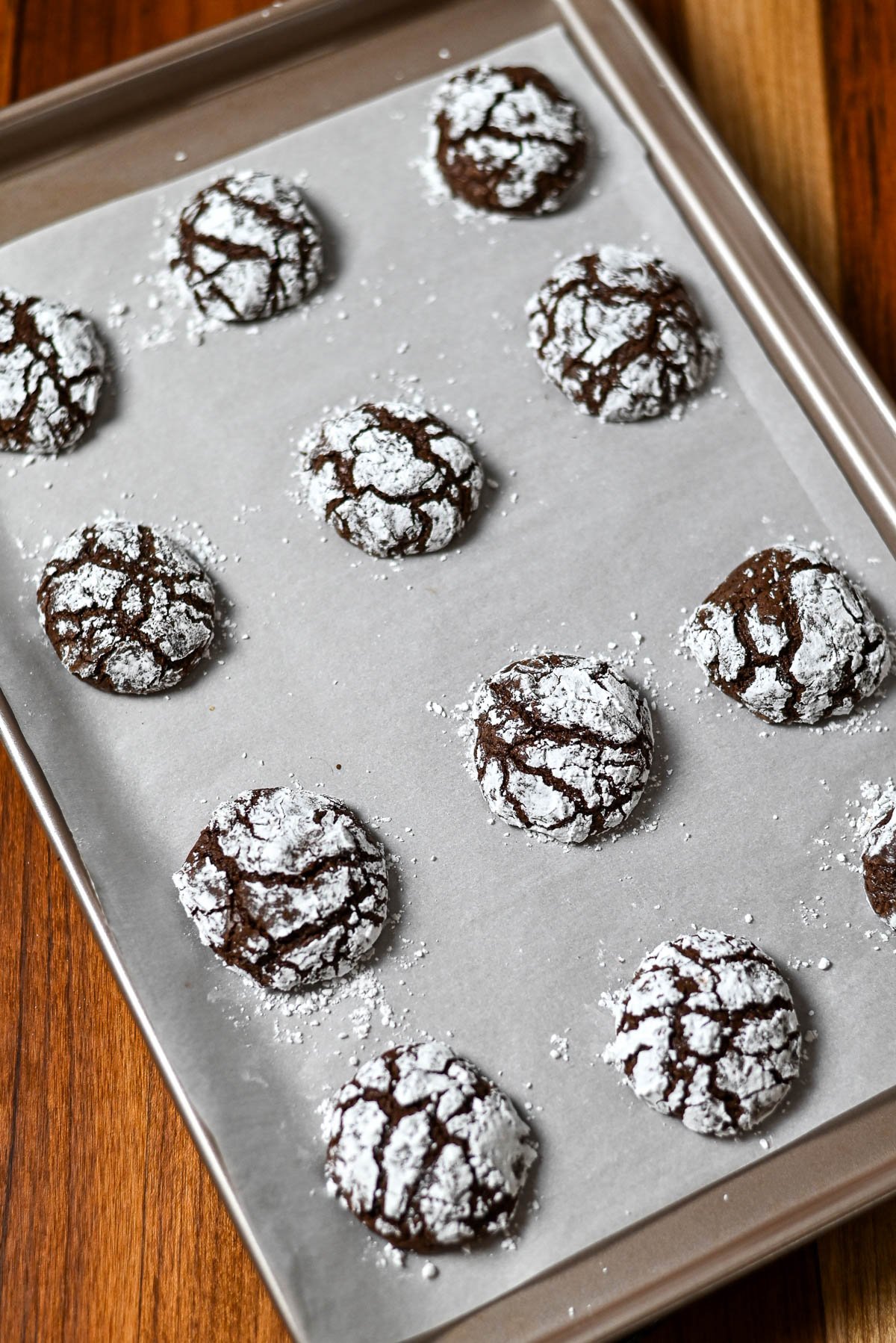 Kiss Crinkle Cookies rest on a parchment-lined baking sheet, dusted with powdered sugar-these chocolate crinkles are irresistibly soft and fudgy.