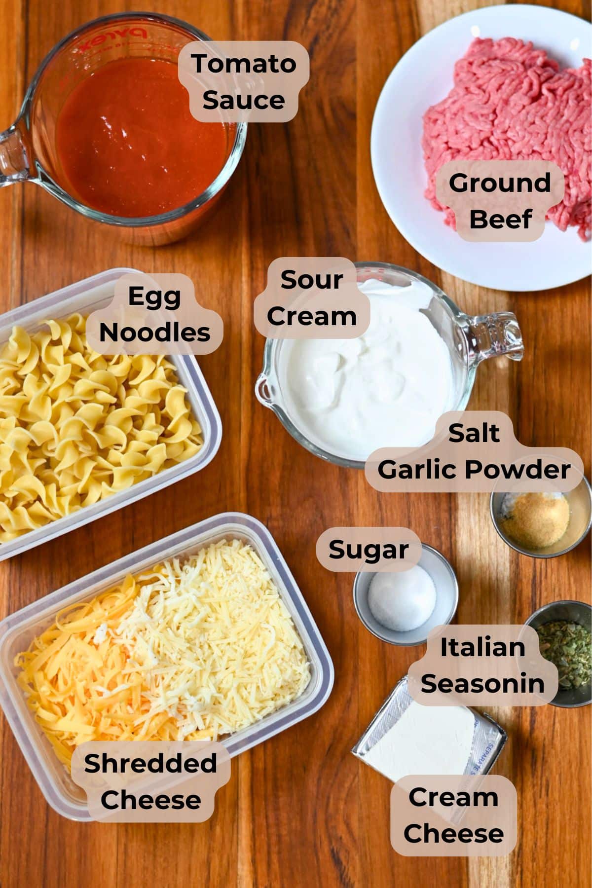 Egg noodles, shredded cheese, tomato sauce, ground beef, sour cream, cream cheese, and seasonings on a table.