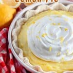 A creamy Sour Cream Lemon Pie topped with whipped cream and lemon zest in a white dish next to lemons and a red checkered cloth.