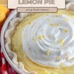 A sour cream lemon pie with whipped topping and lemon zest in a pie dish, next to fresh lemons and a red checkered cloth.