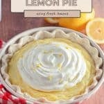 A classic lemon cream pie topped with whipped cream and lemon zest sits on a red-checkered cloth beside fresh lemons.