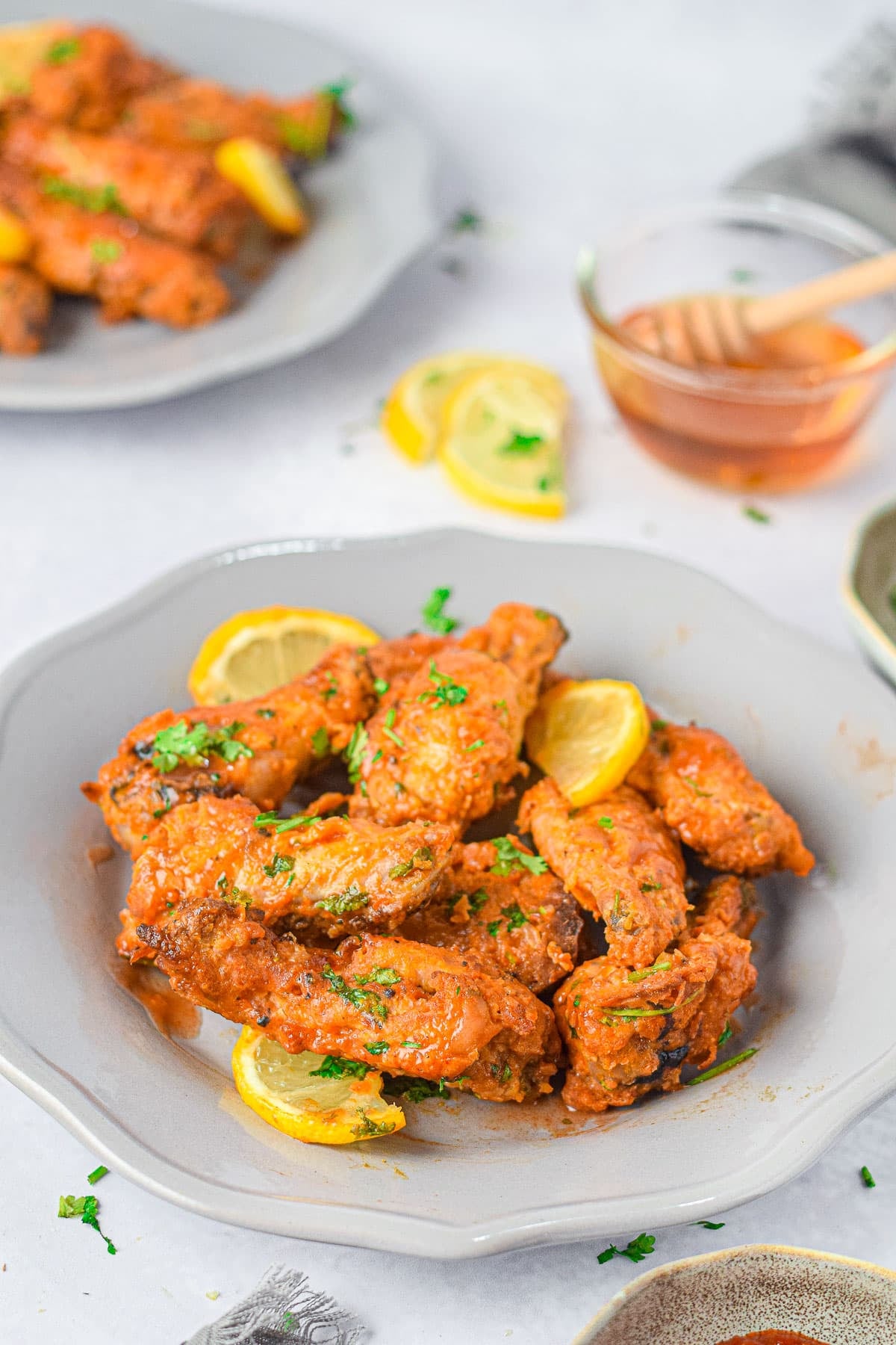 A plate of saucy Air Fryer Honey Lime Chicken Wings garnished with lemon slices and herbs, with a bowl of honey in the background.