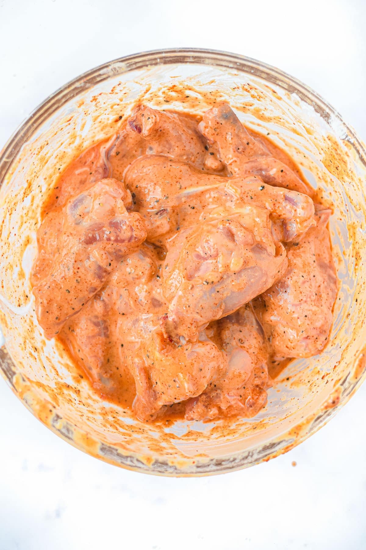 Raw chicken pieces coated in a reddish-orange marinade, ready to become delicious Air Fryer Honey Lime Chicken Wings, in a glass bowl on a white surface.