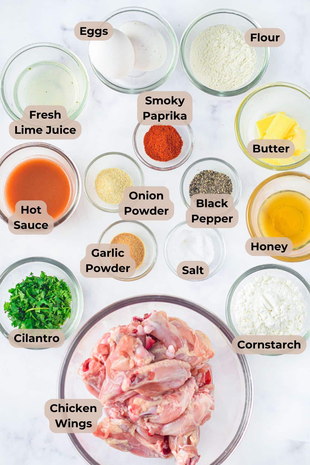Raw chicken wings in a bowl surrounded by labeled bowls of spices, sauces, flour, butter, eggs, and herbs.
