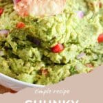 A hand dips a chip into chunky guacamole, filled with tomato and onion pieces, served in a white bowl.