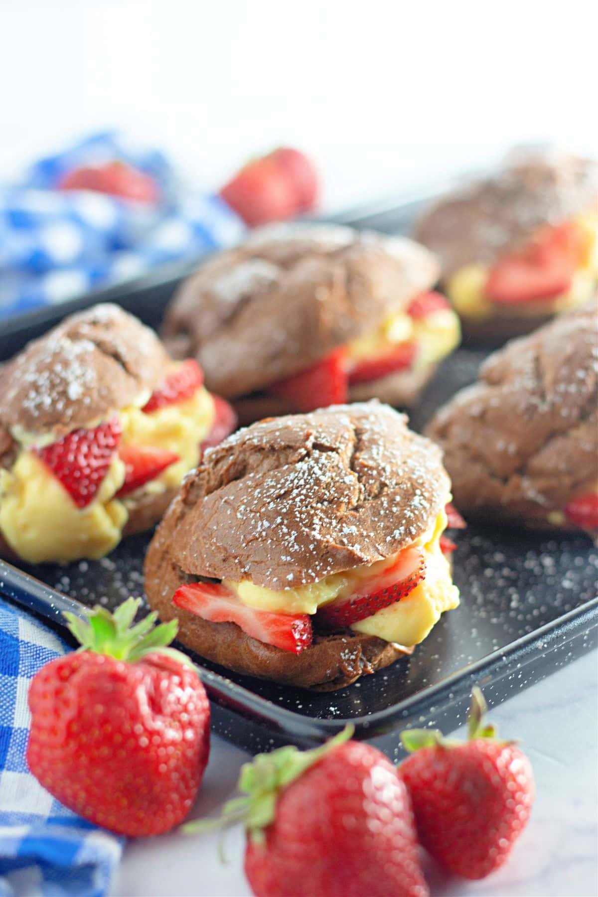 A black baking tray with chocolate cream puffs filled with pudding and strawberries. 