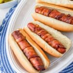 Hot dogs wrapped in crispy bacon in a bun on a white plate.