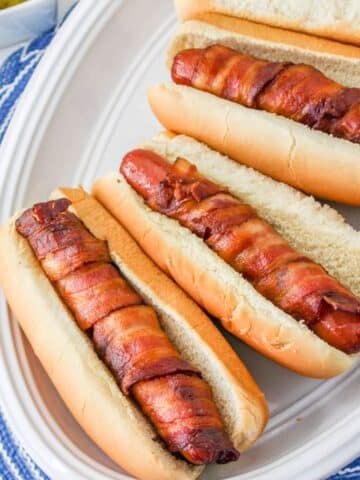 Hot dogs wrapped in crispy bacon in a bun on a white plate.
