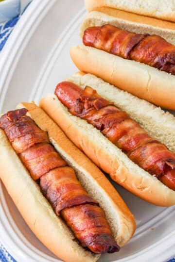 Hot dogs wrapped in crispy bacon in a bun on a white plate.