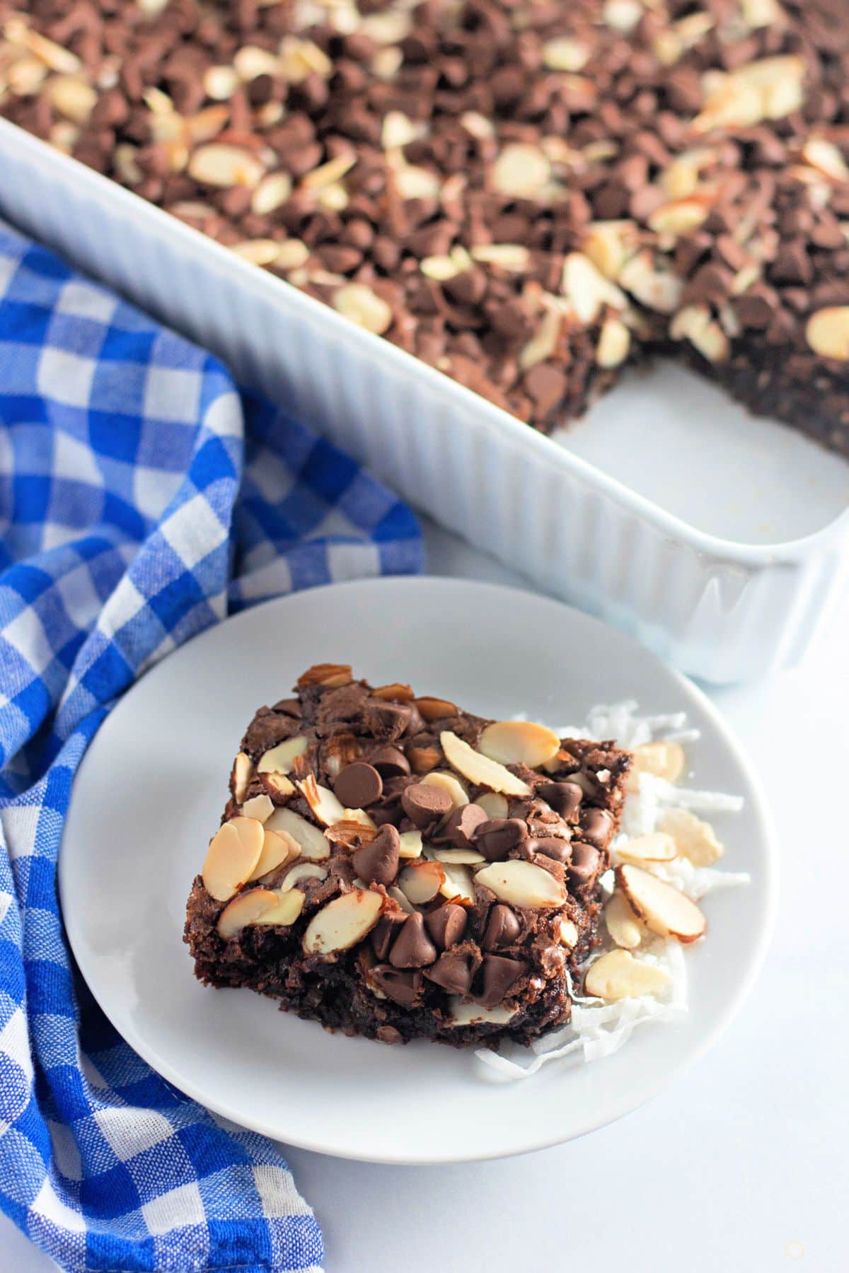 A chocolate chip almond bar on a white plate, with a pan of bars and blue-checkered cloth beside it.