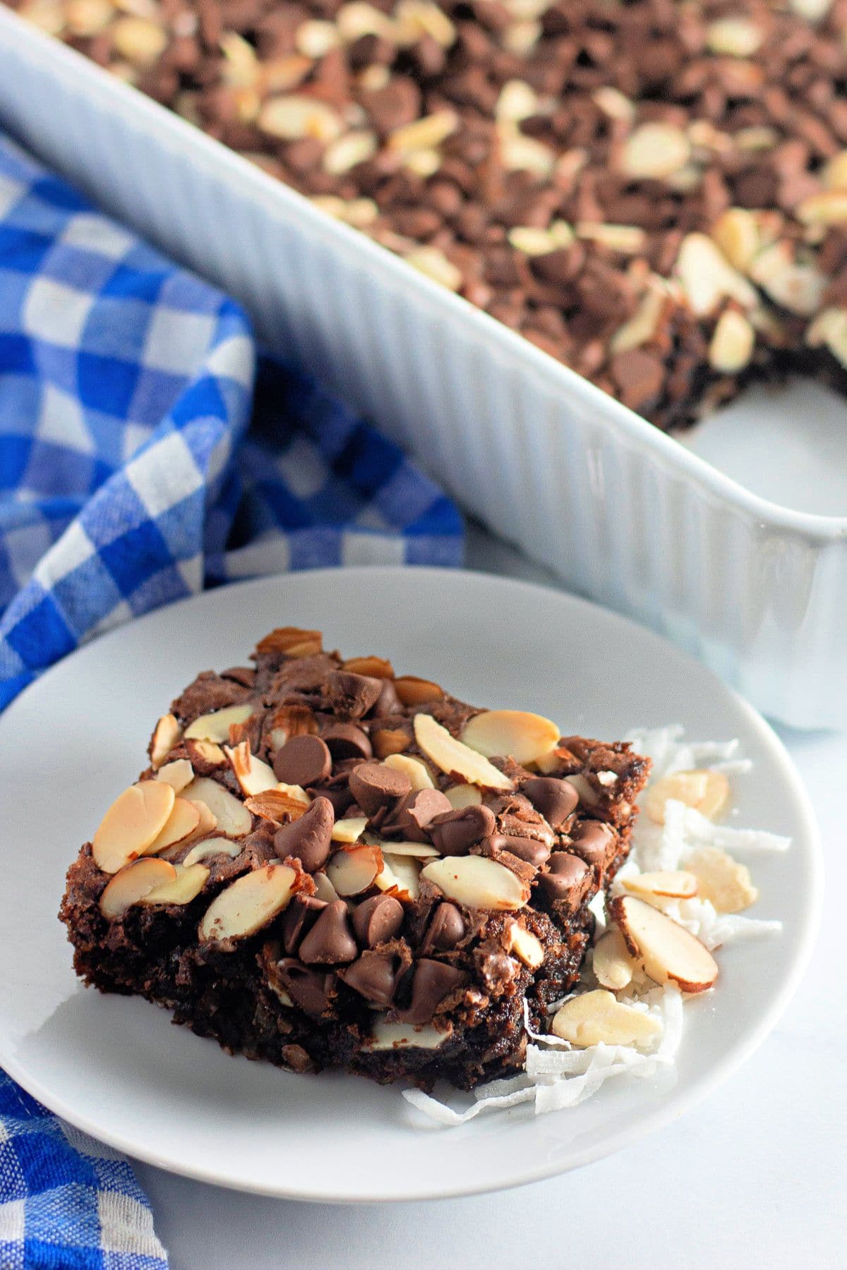 An Easy Coconut Almond Chocolate Brownie dessert bar topped with sliced almonds and chocolate chips sits beautifully on a white plate, with a baking dish behind.
