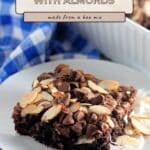 A plate features a rich coconut brownie with almond slices and chocolate chips, an irresistible example of Easy Coconut Almond Chocolate Brownies, with a baking pan and blue checked cloth in the background.