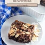 An Easy Coconut Almond Chocolate Brownie topped with chocolate chips and almonds sits on a white plate, next to a blue checked cloth.