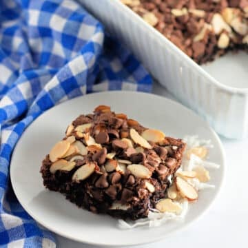 A brownie topped with chocolate chips and sliced almonds on a white plate beside a blue checkered cloth.