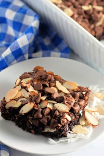A brownie topped with chocolate chips and sliced almonds on a white plate beside a blue checkered cloth.