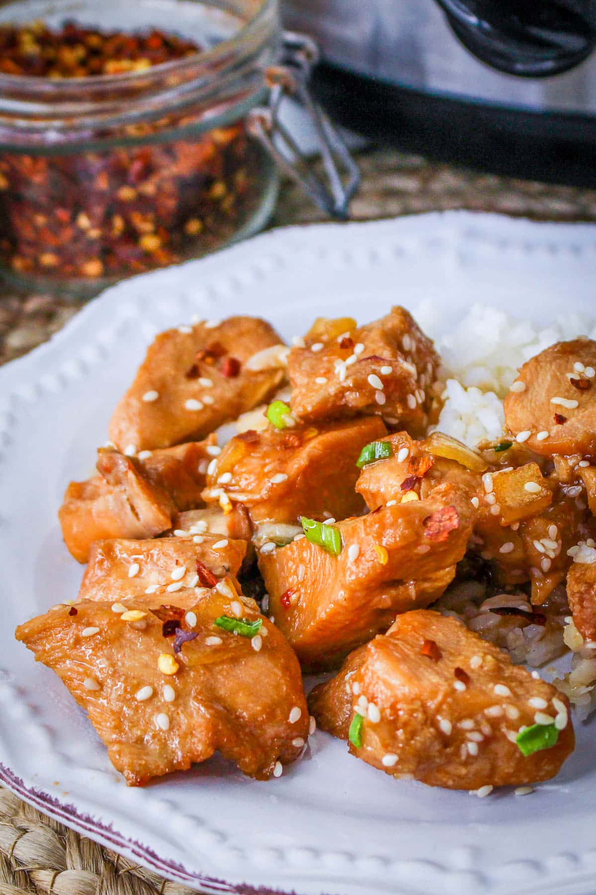 Plate of Slow Cooker Sesame Chicken with rice, garnished with green onions and red pepper flakes, jar of spices in background.