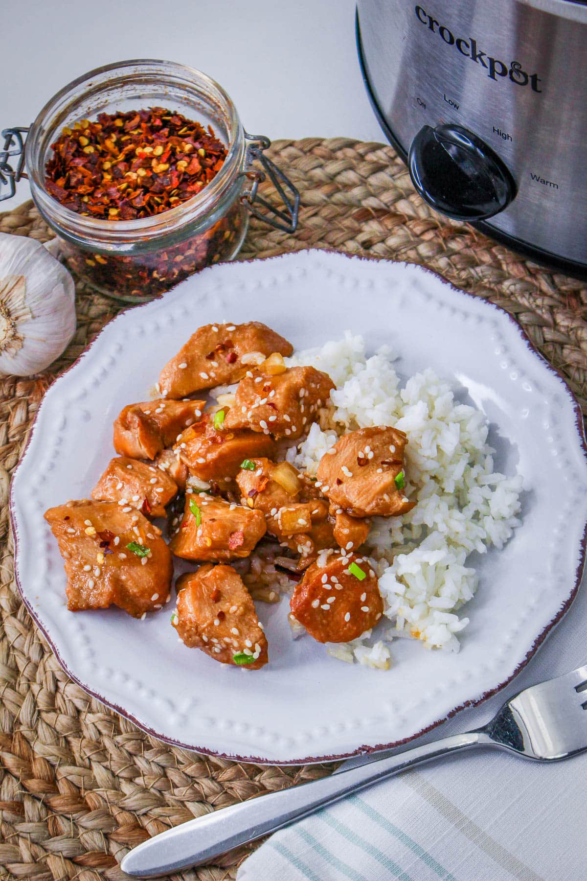 A plate of Slow Cooker Sesame Chicken with sauce and white rice sits beside a slow cooker, aromatic spices, and a fresh garlic bulb.