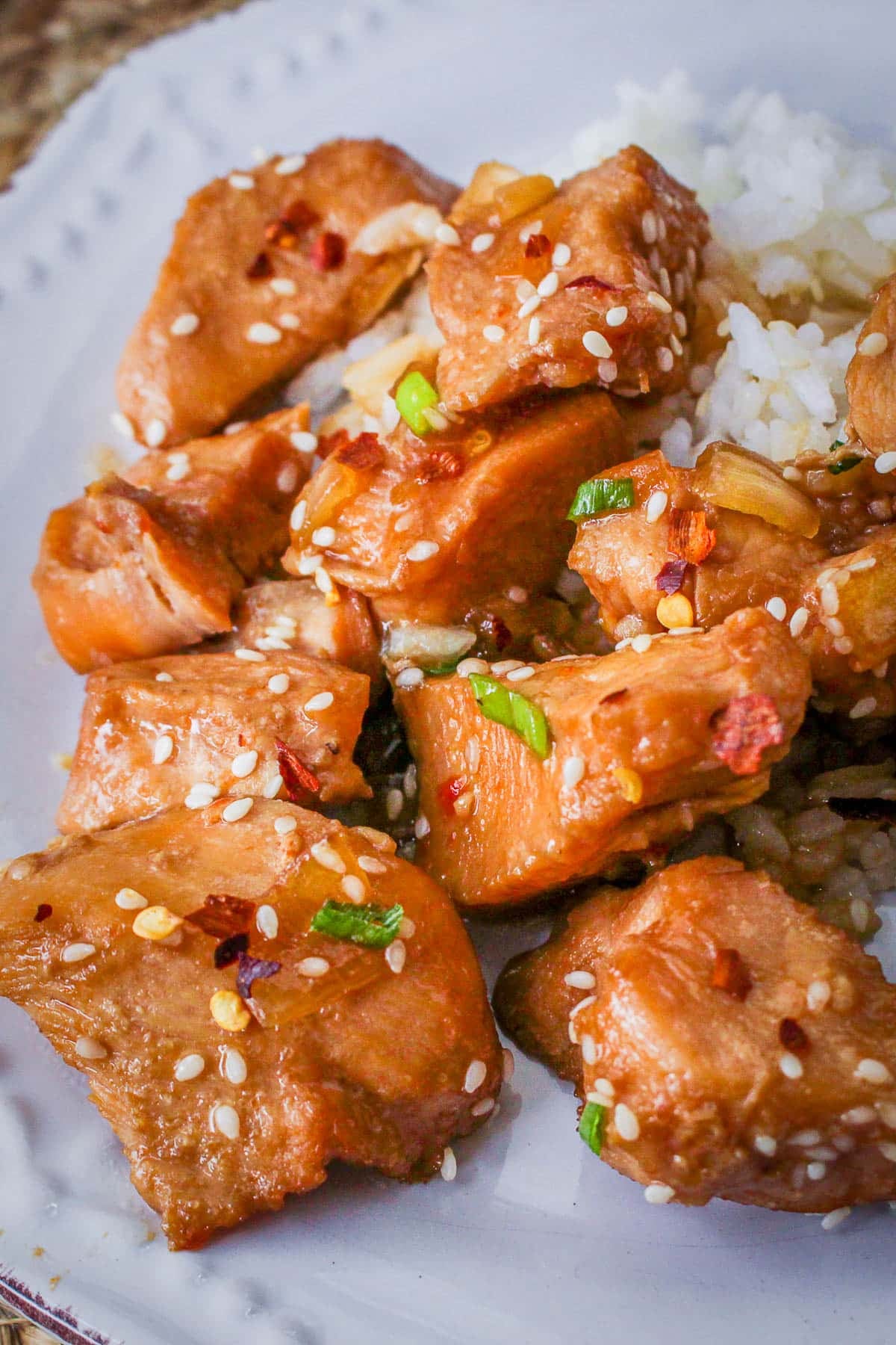 Chunks of glazed Slow Cooker Sesame Chicken with sesame seeds and green onions, served with white rice on a white plate.