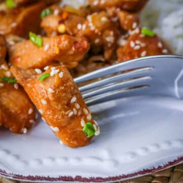 A fork holds a sesame-coated piece of Slow Cooker Sesame Chicken, garnished with green onion, on a white plate.