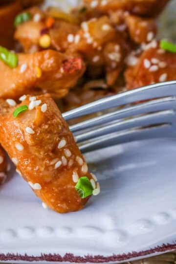 A fork holds a sesame-coated piece of Slow Cooker Sesame Chicken, garnished with green onion, on a white plate.