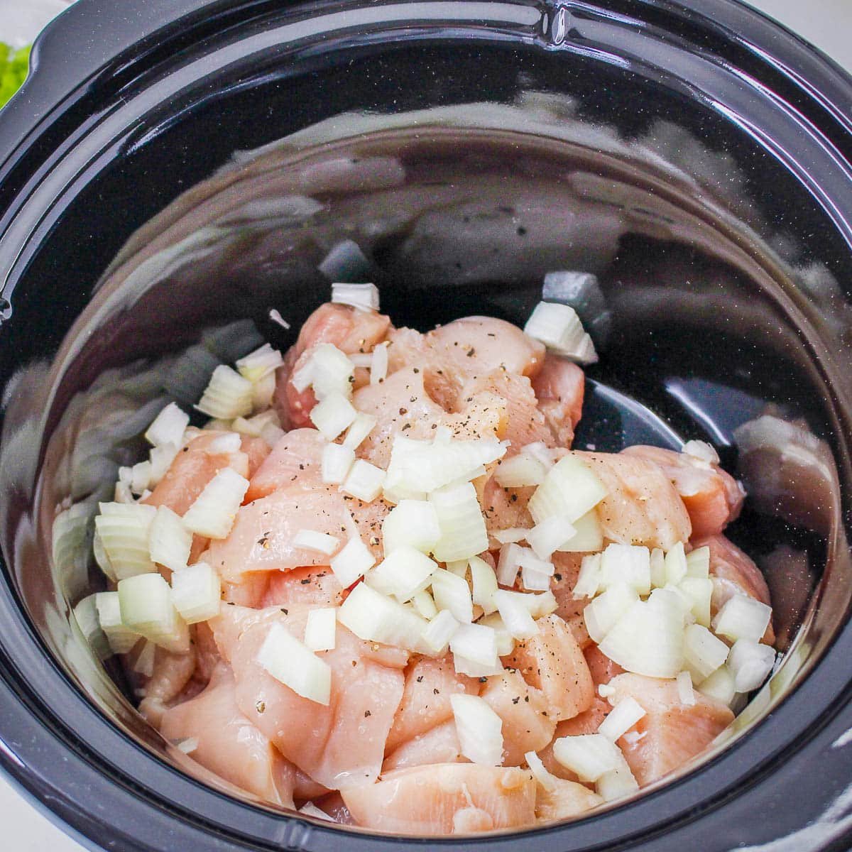 Chopped raw chicken and diced onions in a black slow cooker, ready to be cooked for a delicious Slow Cooker Sesame Chicken.