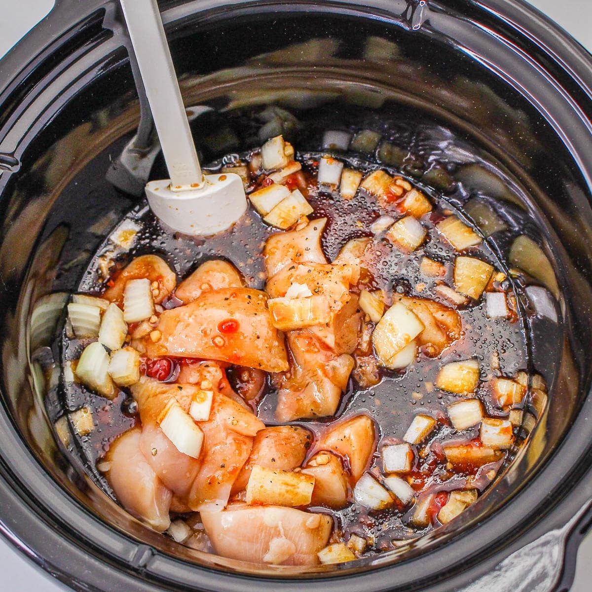 Raw chicken, chopped onions, and sauce in a slow cooker with a spatula-perfect for preparing flavorful Slow Cooker Sesame Chicken.