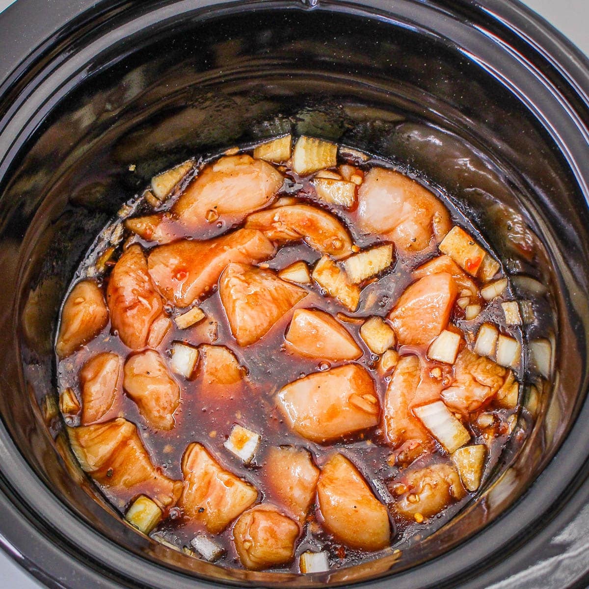 Raw chicken pieces and chopped onions coated in a rich, dark sesame sauce inside a slow cooker, ready to transform into tender Slow Cooker Sesame Chicken.