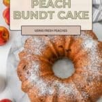 A Peach Bundt Cake dusted with powdered sugar sits on a marble surface, surrounded by fresh peaches, inviting you to enjoy a slice of summer sweetness.