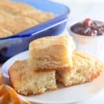 Biscuits stacked on a white plate next to a baking dish a bowl of jam.