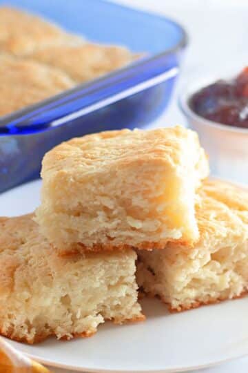 Biscuits stacked on a white plate next to a baking dish a bowl of jam.