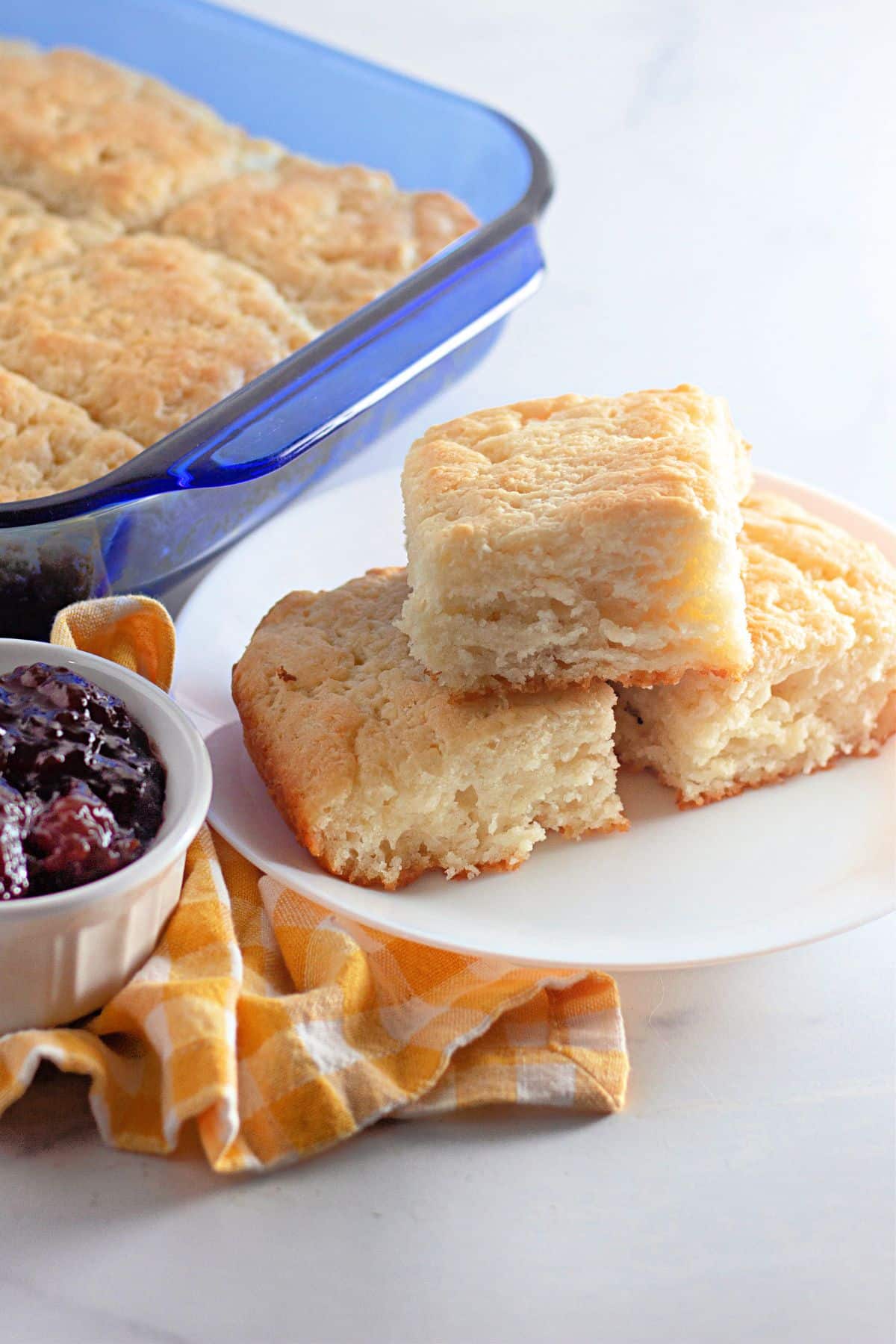 Biscuits stacked on a white plate next to a baking dish a bowl of jam.