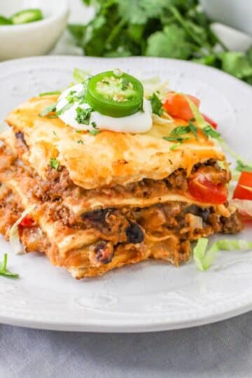 An individual serving of taco lasagna on a white plate next to a fork and taco toppings.