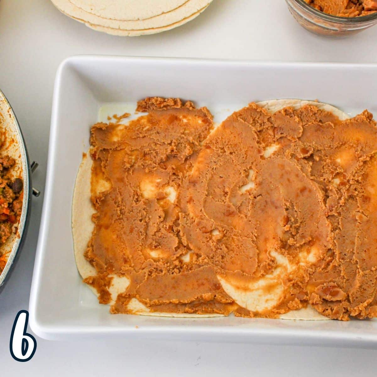 Refried beans spread over a layer of tortillas in a baking dish. 