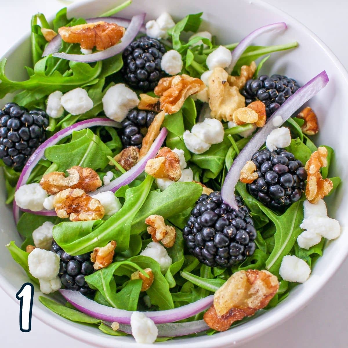 Arugula mixed with walnuts, red onion, goat cheese, and blackberries in a white bowl. 