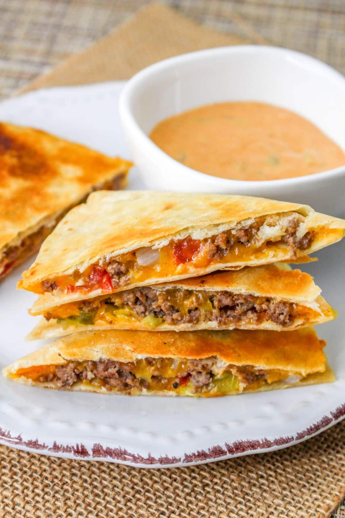 Cheeseburger quesadillas stacked on a white plate next to a bowl of special sauce. 