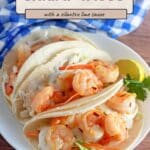 Three shrimp tacos with slaw and cilantro lime sauce on a white plate, served with a lime wedge and a blue checkered napkin.