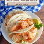 Three Shrimp Tacos with Cilantro Lime Sauce on a plate, garnished with lemon and cilantro, blue napkin beside.
