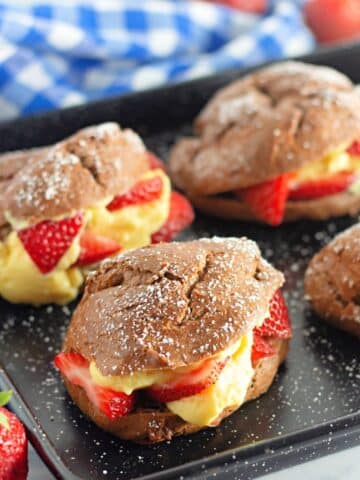 A black baking tray with chocolate cream puffs filled with pudding and strawberries.