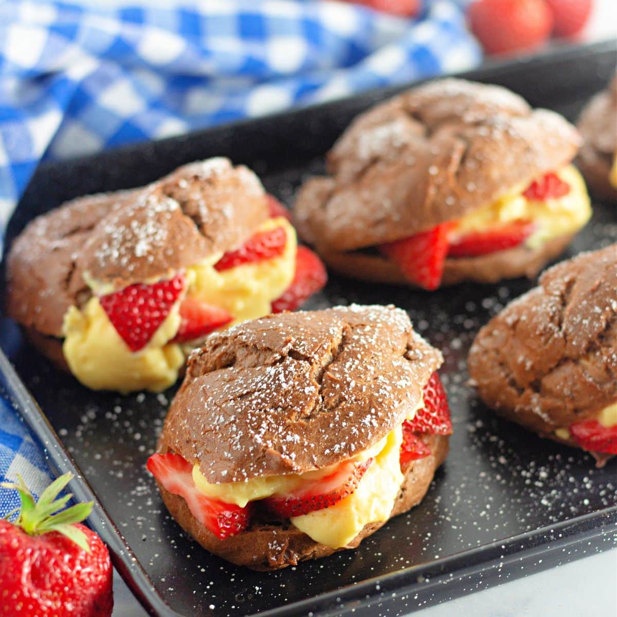 A black baking tray with chocolate cream puffs filled with pudding and strawberries.