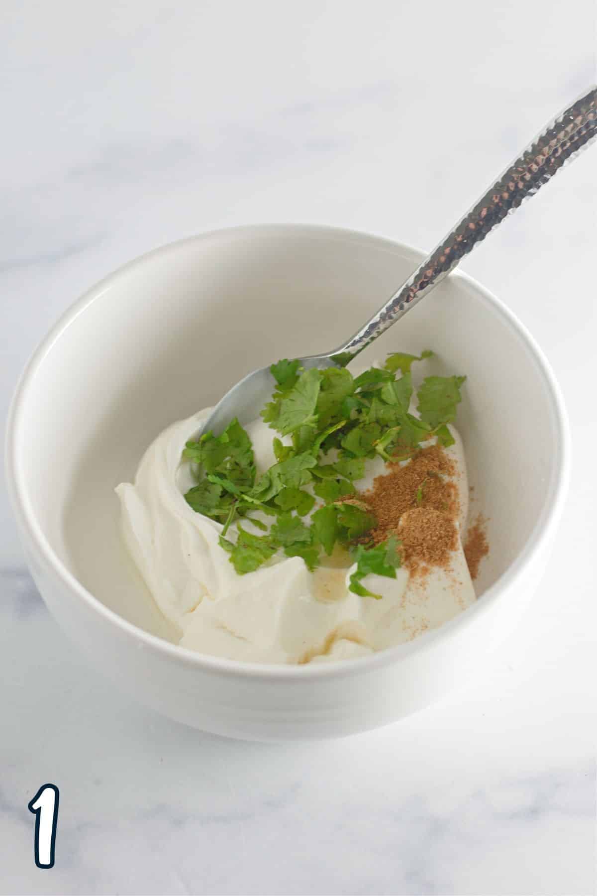 Sour cream, cumin, lime juice, and cilantro in a bowl with a spoon. 