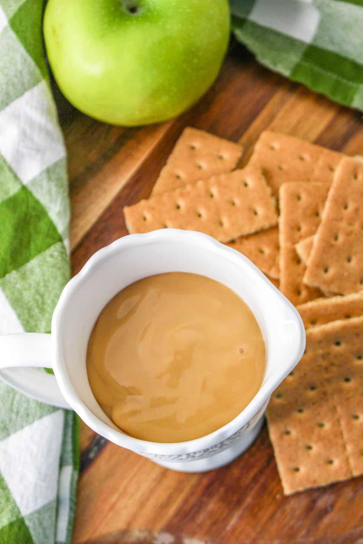 A white cup filled with Dulce de Leche next to green apples and graham crackers. 