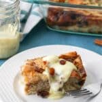 A piece of bread pudding on a white plate next to a jar of vanilla sauce.