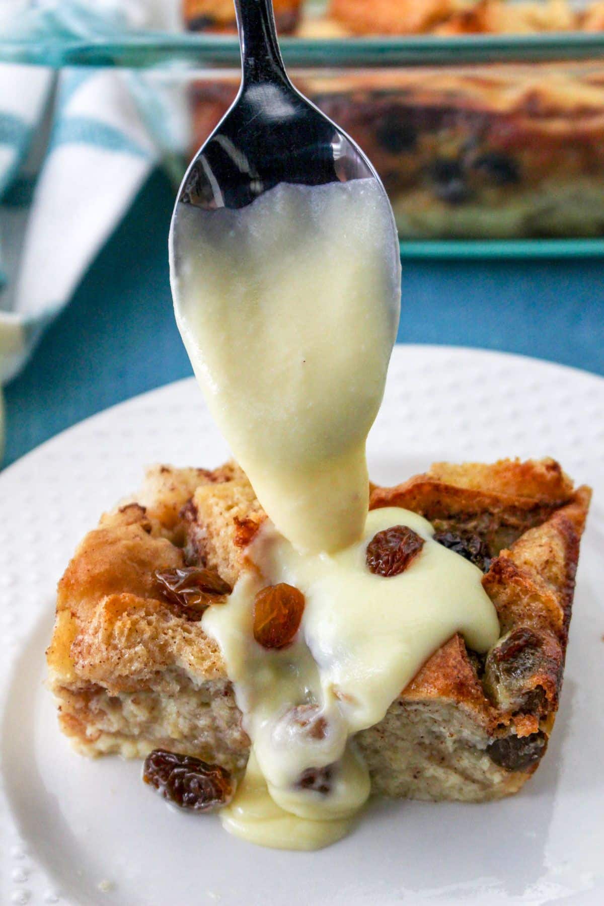 A spoon dripping vanilla sauce over a piece of bread pudding on a white plate. 
