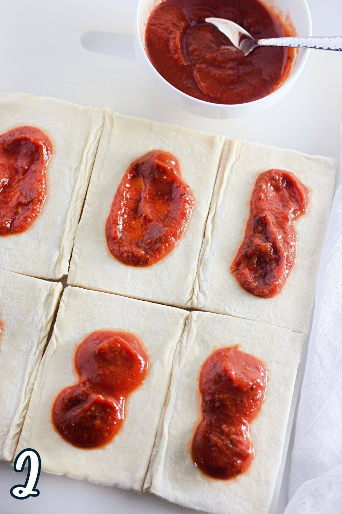 A sheet of puff pastry cut into 6 rectangles and pizza sauce in the center of each one. 