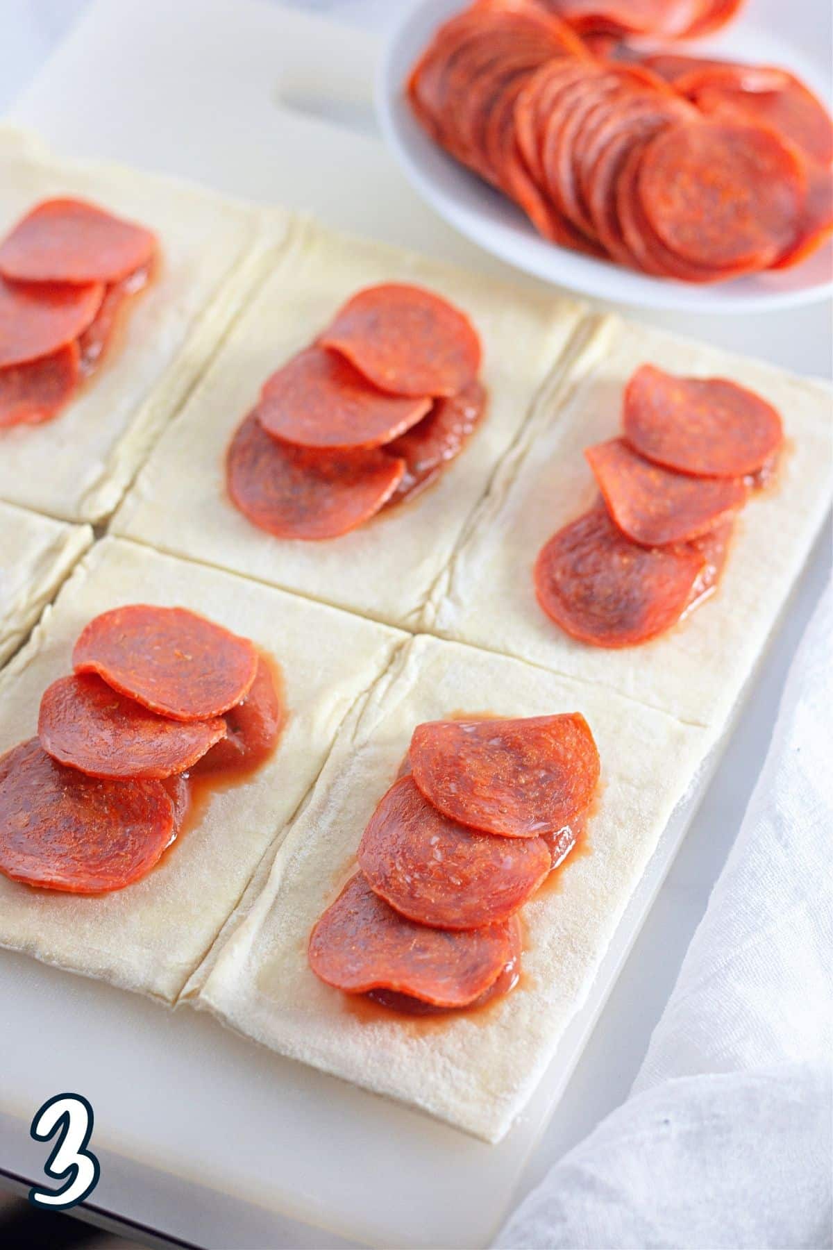Sliced pepperoni laid over pizza sauce on rectangles of puff pastry. 