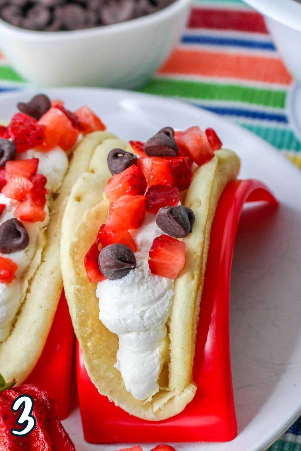 A red taco holders holding a pancake filled with cheesecake mousse and fresh strawberries with chocolate chips. 