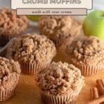 Apple Cinnamon Muffins on a wooden surface with cinnamon sticks and a green apple in the background. Each muffin boasts a golden crumb topping, highlighting the comforting flavors of apple and warm spice in every bite.